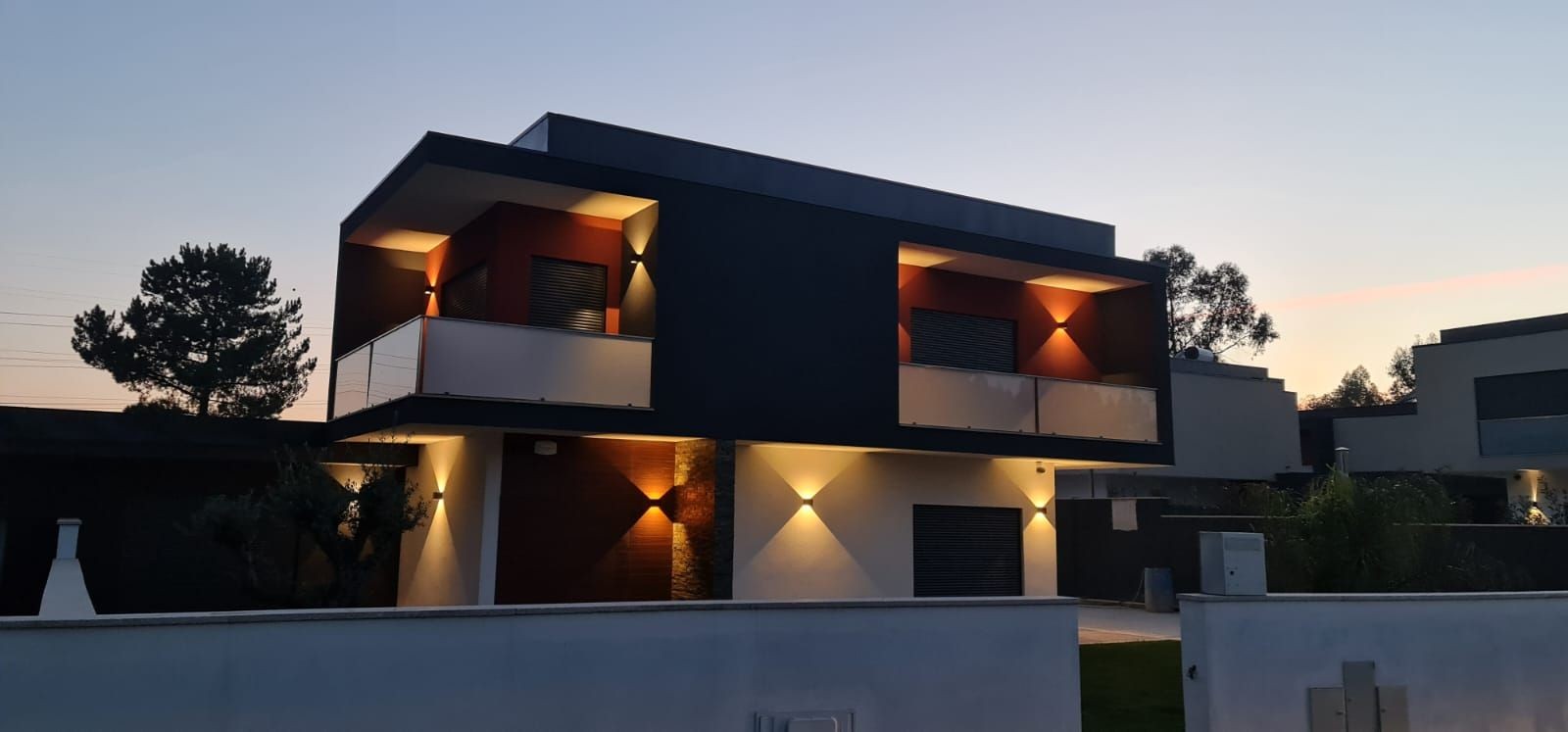 Modern two-story house with evening lighting and a balcony, set against a dusky sky.