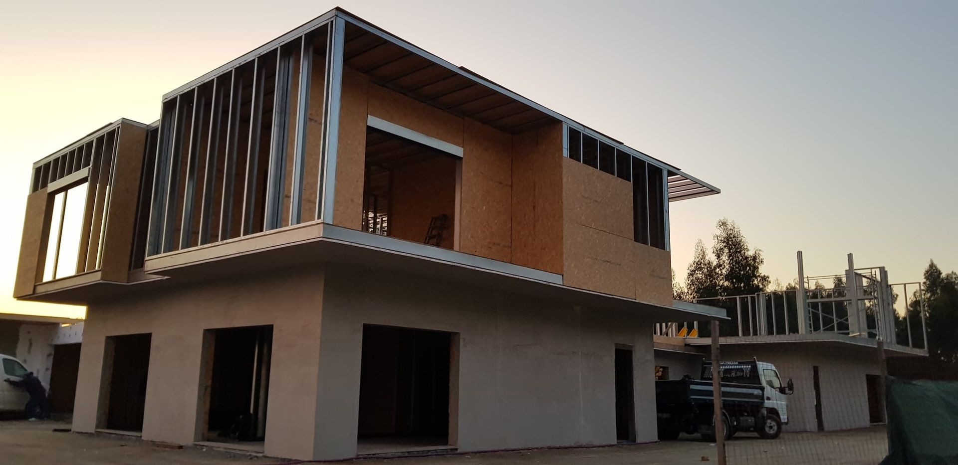 Modern two-story building under construction with metal and wood framework, surrounded by trees at sunset.